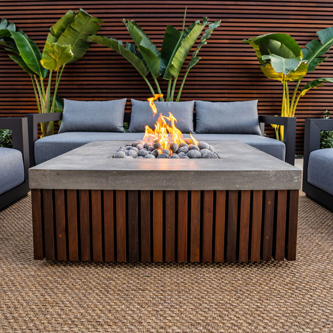 The Manchester fire table in a carpeted outdoor seating area with gray-blue couches and large green plants.