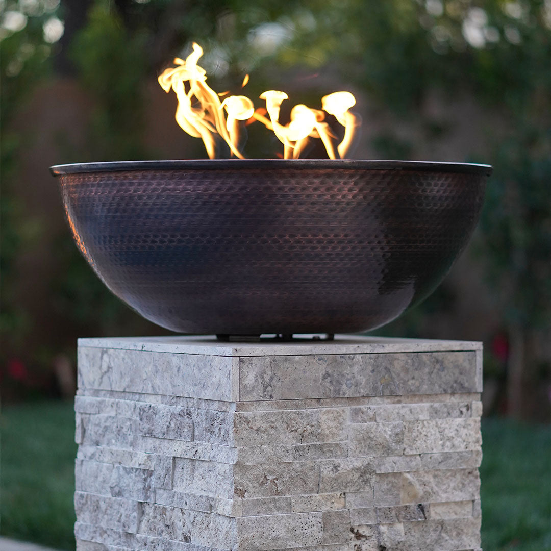 The Sedona Fire Bowl in hammered copper by The Outdoor Plus installed on a pedestal and lit.